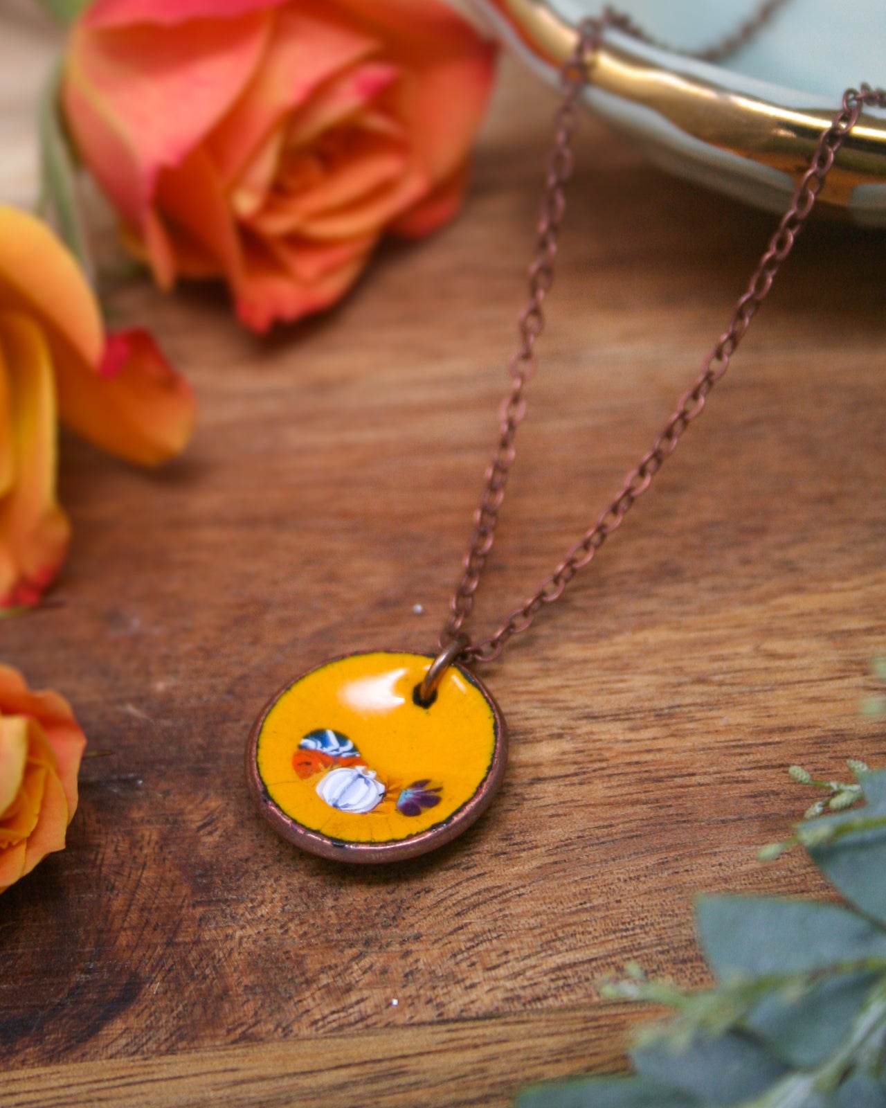 A yellow pendant necklace with a circular design and small objects is displayed on a wooden surface, surrounded by orange and red roses.