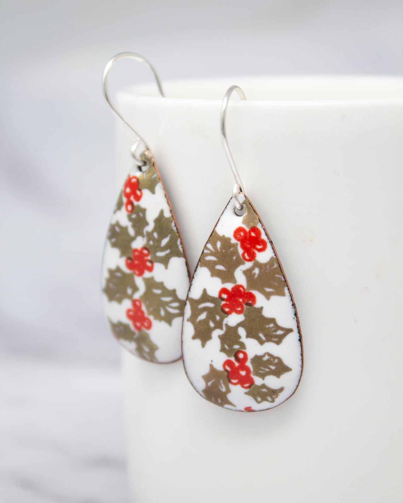 A pair of earrings with a floral pattern, featuring red berries and leaves, are displayed on a white surface.