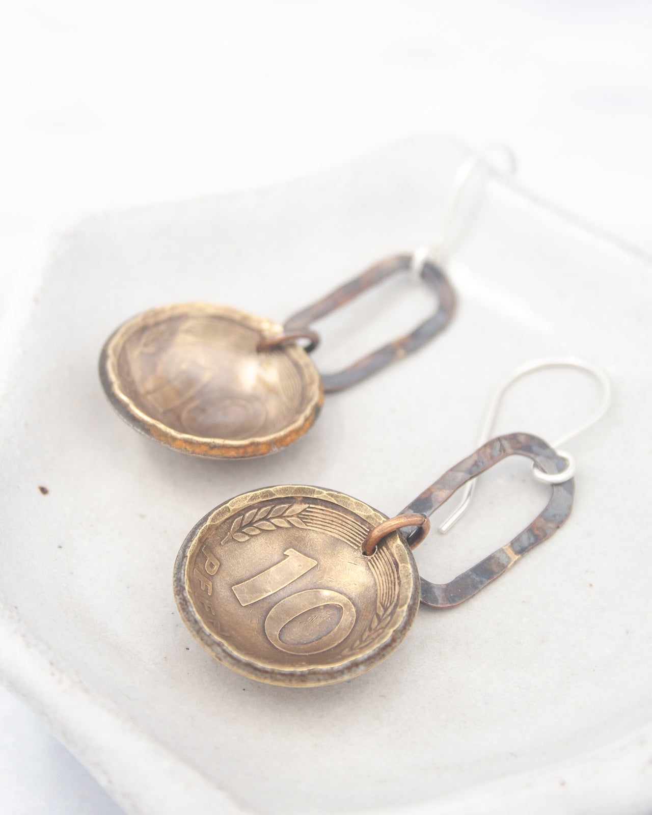A pair of earrings made of metal, featuring a design that resembles a coin. The earrings have a metallic appearance and are displayed on a white surface.