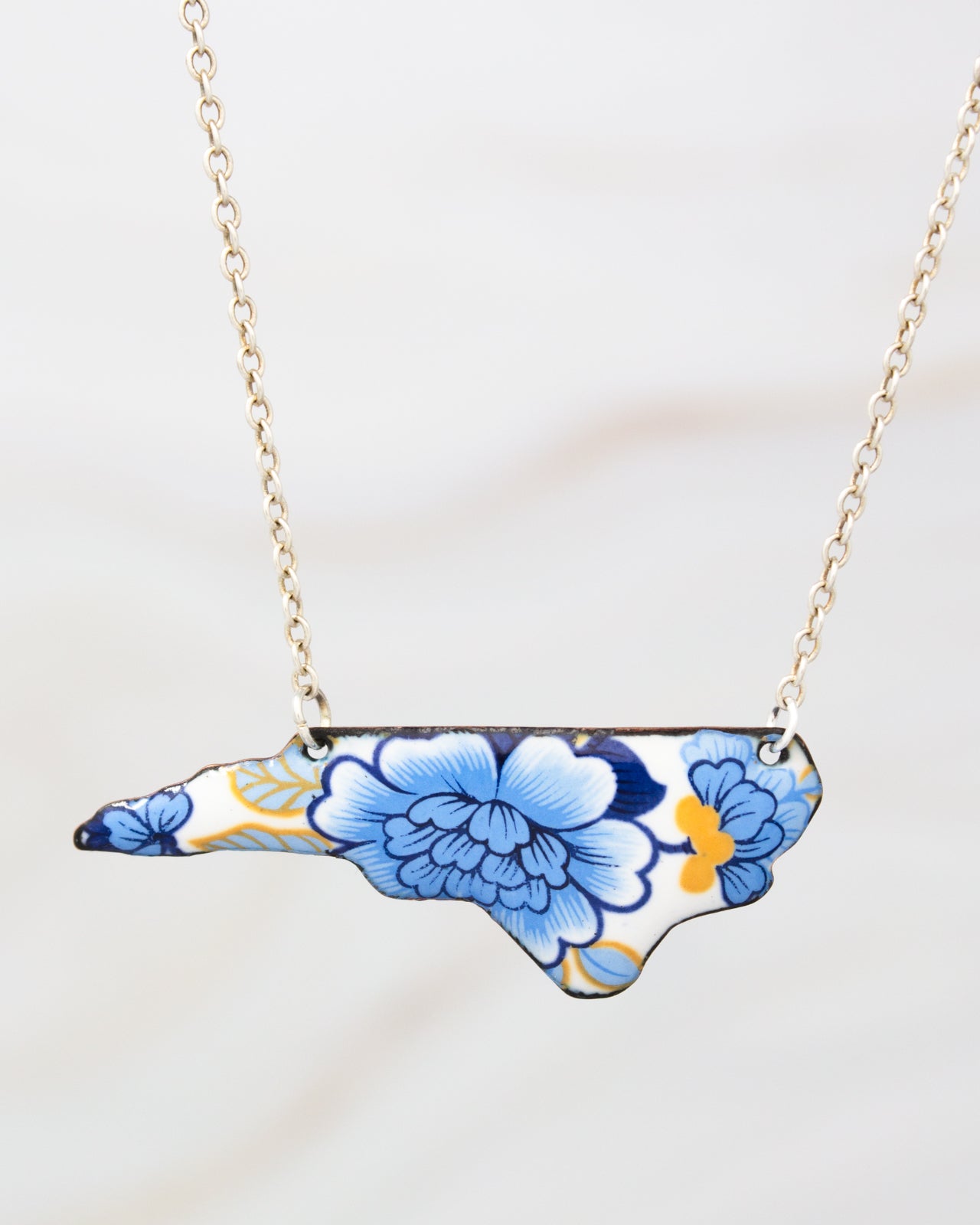 A necklace with a pendant in the shape of the state of North Carolina, featuring a blue and white floral design.