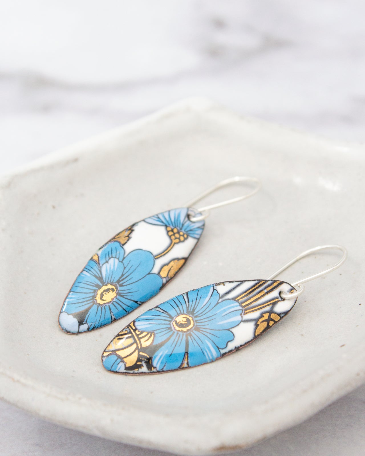 A pair of earrings with blue and gold floral designs, placed on a white plate against a blurred background.