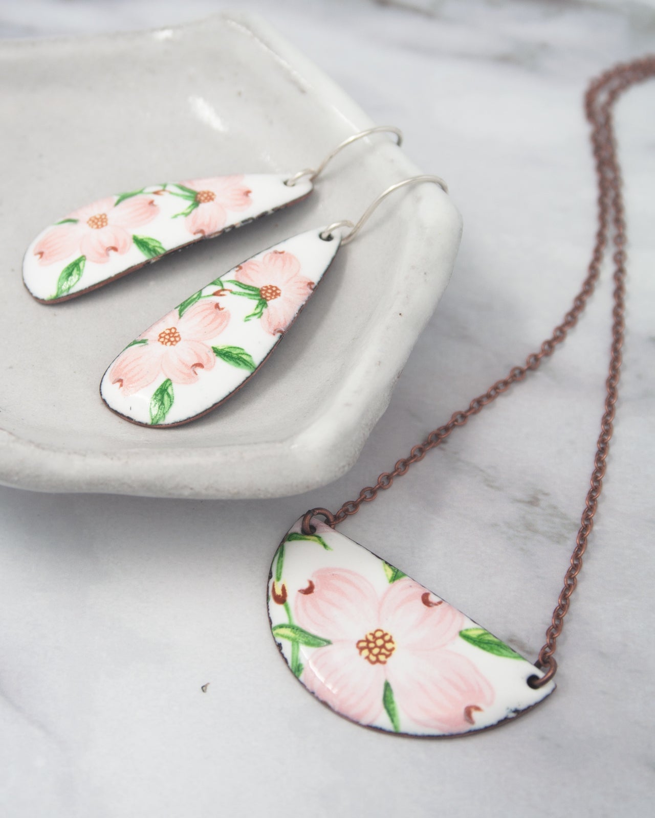 a pair of earrings and a necklace with floral designs, including pink flowers and green leaves, resting on a white surface.