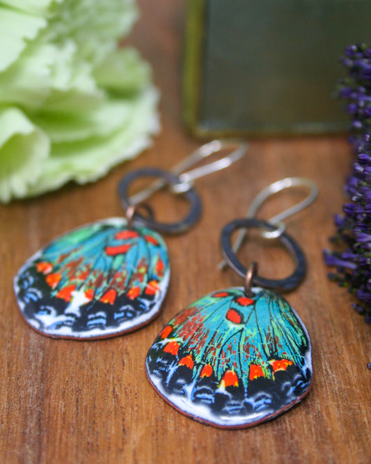 a pair of colorful earrings with butterfly designs, placed on a wooden surface alongside some purple flowers.