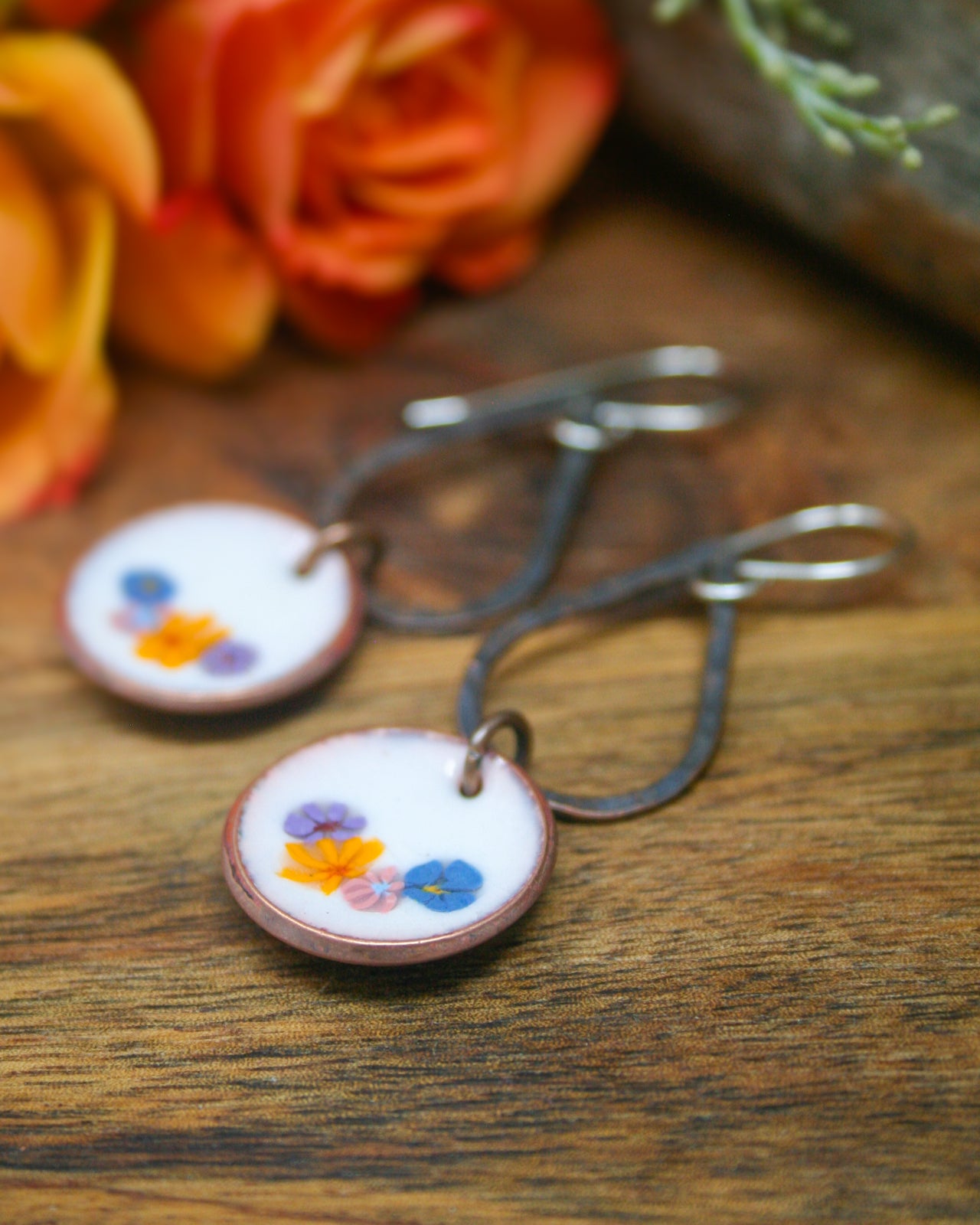 A pair of round earrings with colorful flower designs on a wooden surface, with a blurred background of orange and yellow flowers.