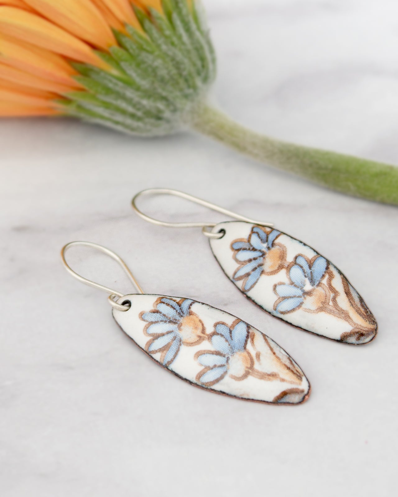 A pair of earrings with blue and brown floral designs, placed on a white surface next to a single orange flower.