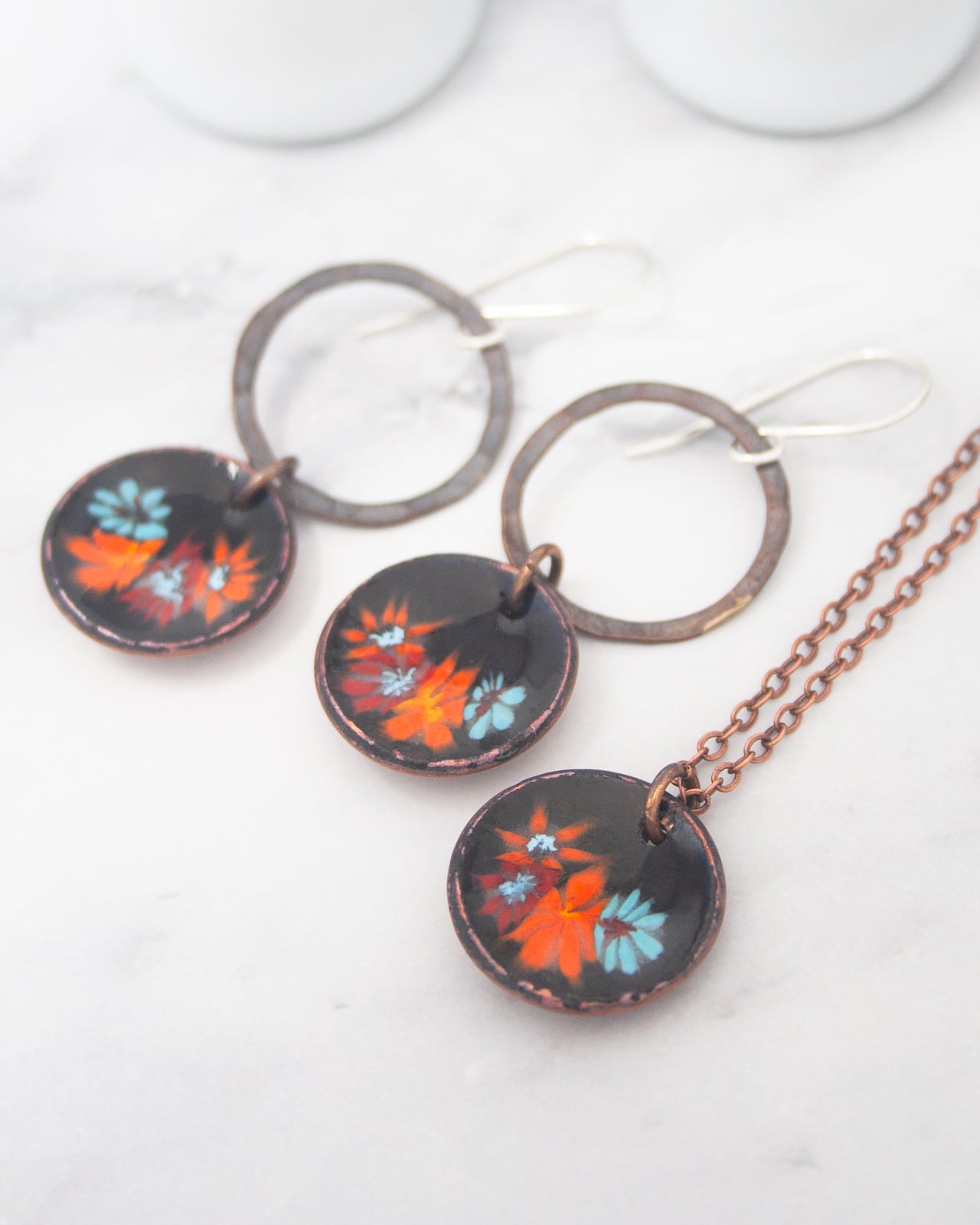 a pair of earrings and a necklace with circular pendants, each adorned with a floral design in shades of orange and blue.