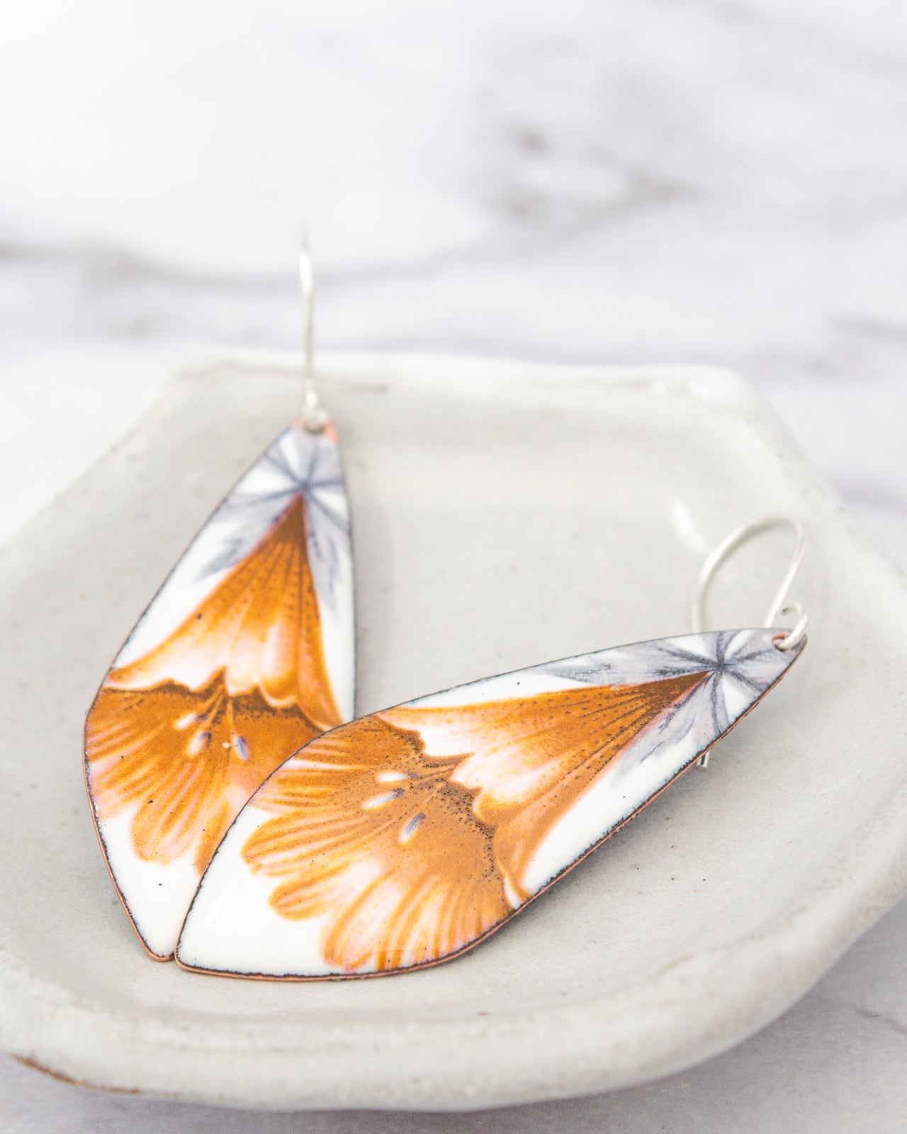 A pair of earrings with a floral design, featuring orange and white colors, are displayed on a white plate against a blurred marble background.