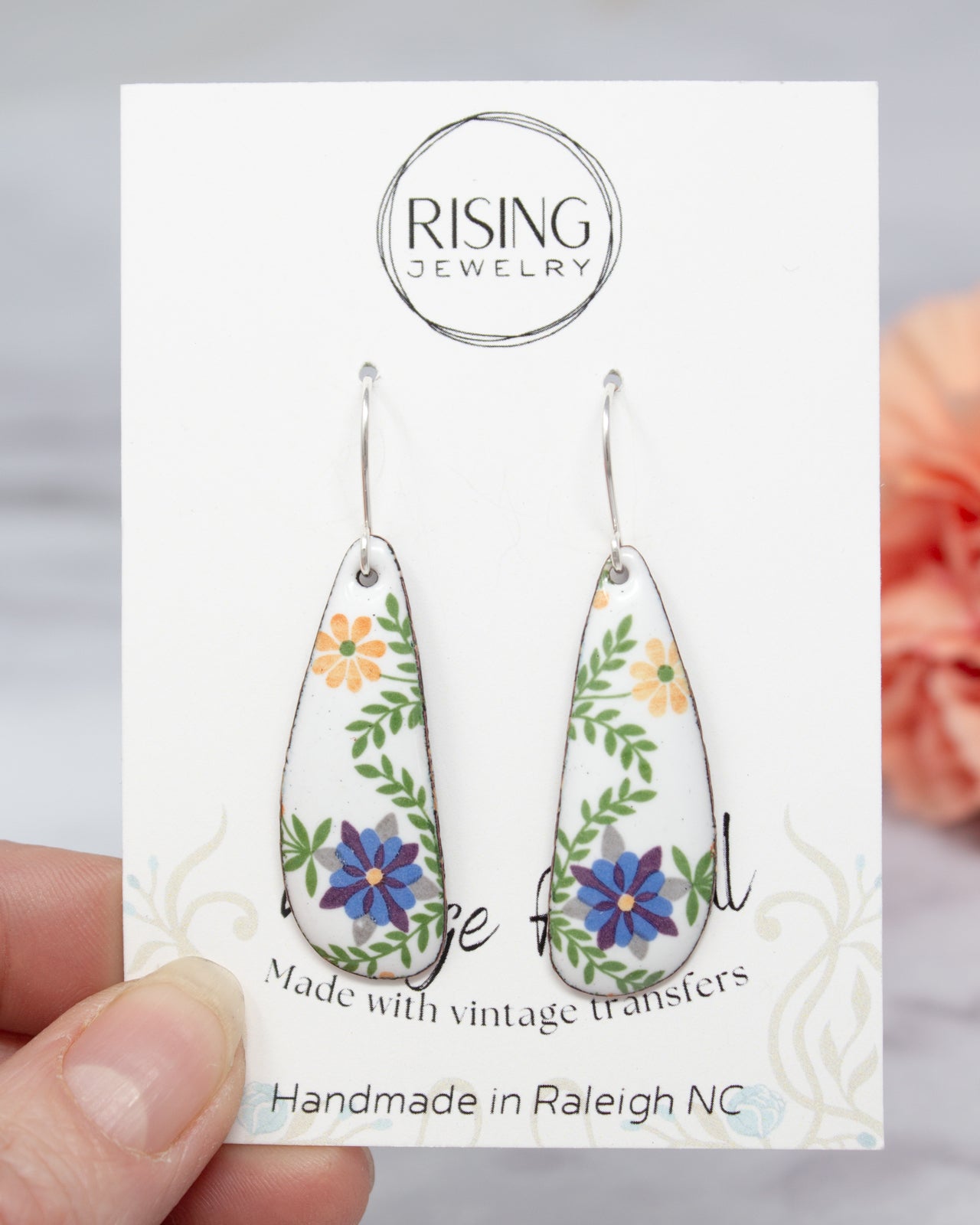A hand holding a pair of earrings with a floral design, against a blurred background.