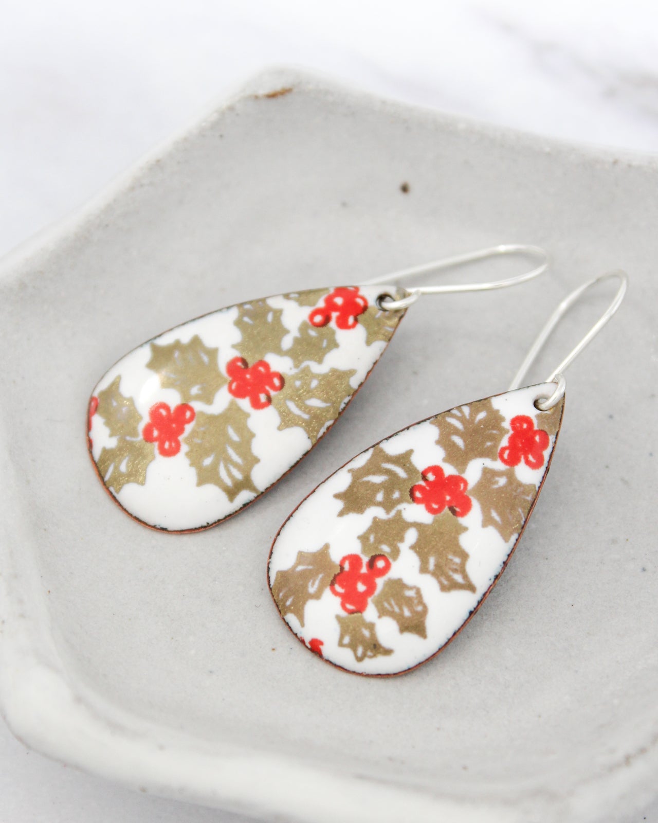 A pair of earrings with a floral design, featuring red berries and gold leaves, resting on a white surface.