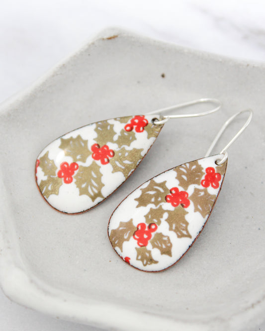 A pair of earrings with a floral design, featuring red berries and gold leaves, resting on a white surface.