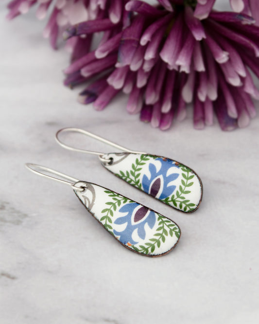 A pair of earrings with a floral design rests on a marble surface, with a vibrant purple flower in the background.