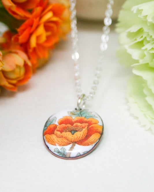 A necklace with a pendant featuring an orange flower design is displayed against a backdrop of orange and green flowers.