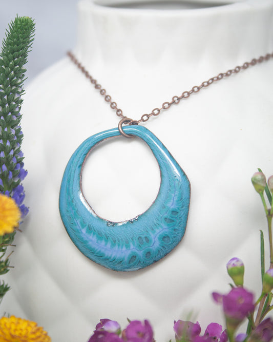 A turquoise pendant necklace with a circular cutout in the center, hanging from a chain. The necklace is displayed against a white background, with colorful flowers visible in the foreground.