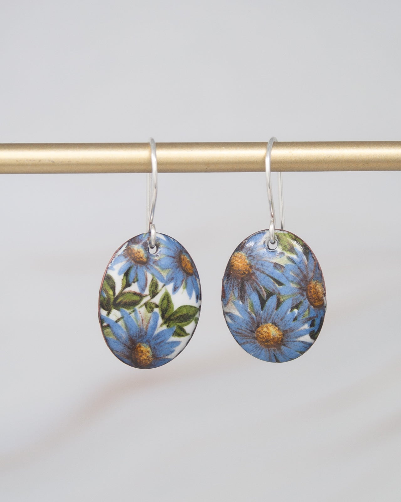 A pair of earrings with blue and green floral designs hanging from a gold-colored metal bar.