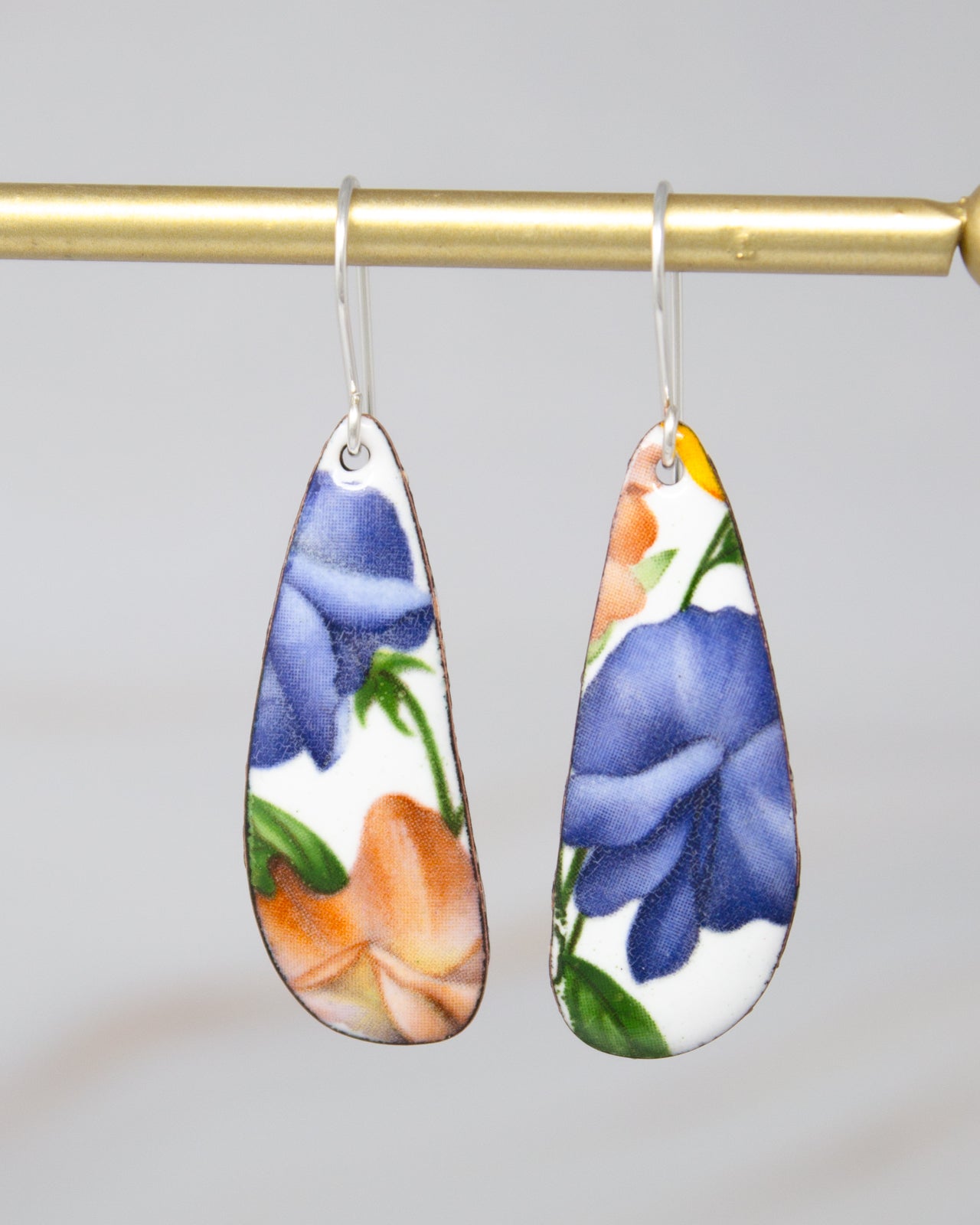 a pair of earrings hanging from a gold-colored metal bar. The earrings have a floral design, with one earring featuring a blue flower and the other earring featuring an orange flower.
