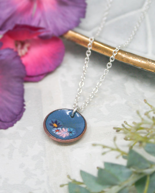 A necklace with a blue pendant featuring a floral design is displayed on a white surface, surrounded by purple and green flowers.