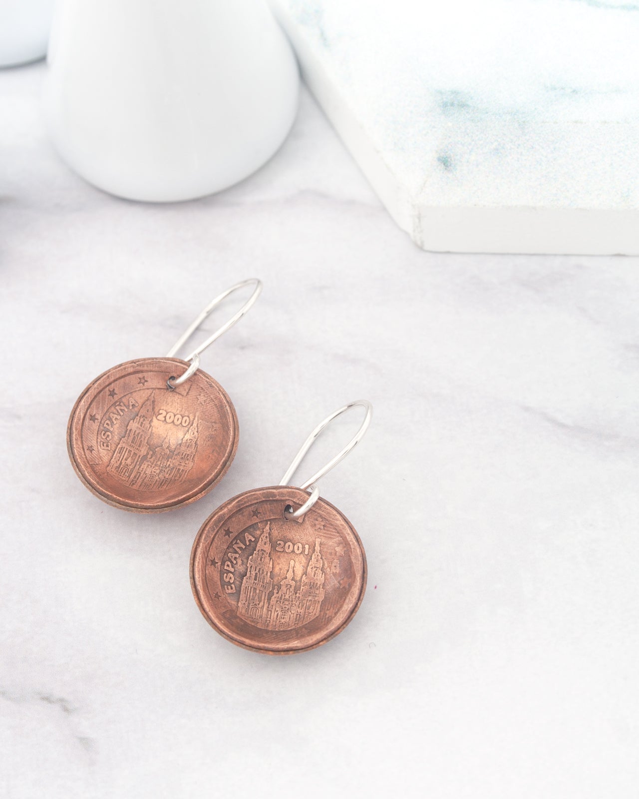 a pair of earrings made from copper-colored metal, featuring a design that resembles a building or monument. The earrings are placed on a white marble surface, with a white ceramic object and a white rectangular object in the background.