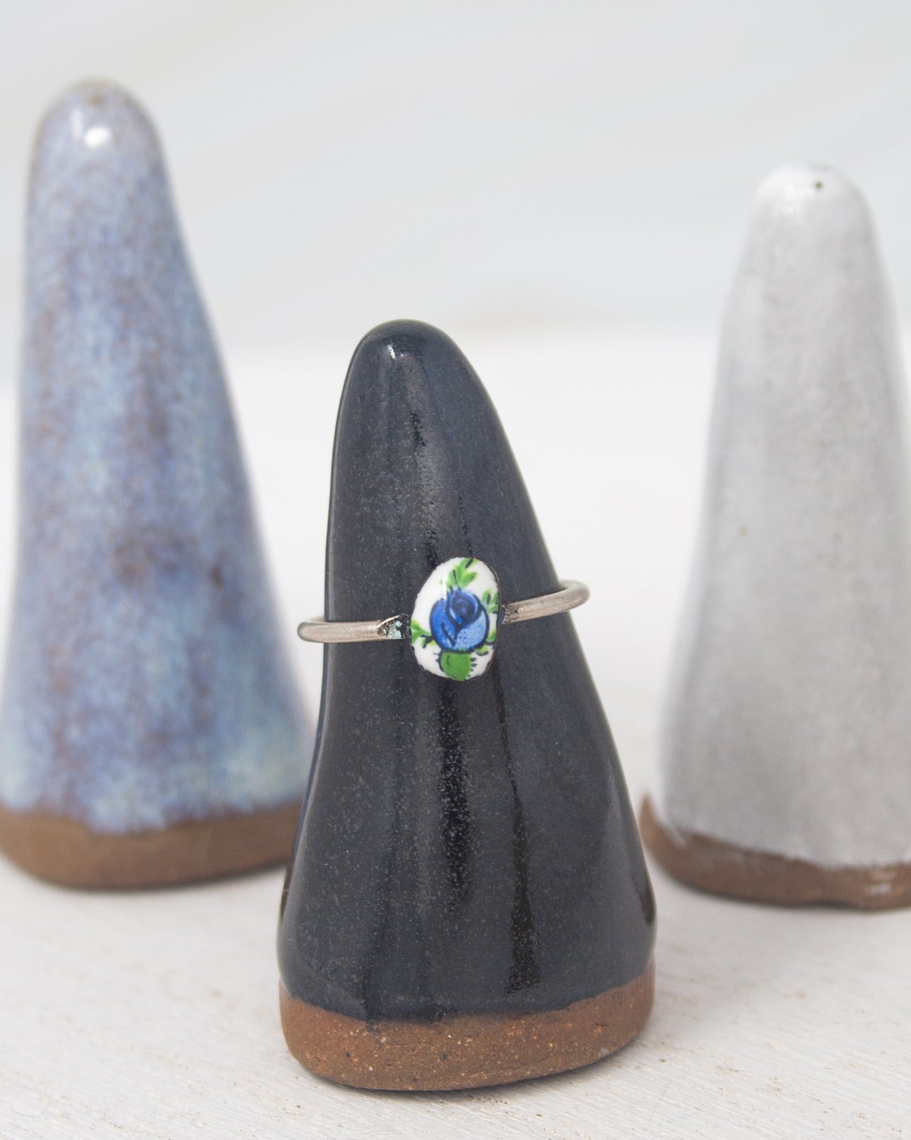 A black ceramic ring stand holding a silver ring with a blue flower design, placed between two white ceramic cone-shaped stands.