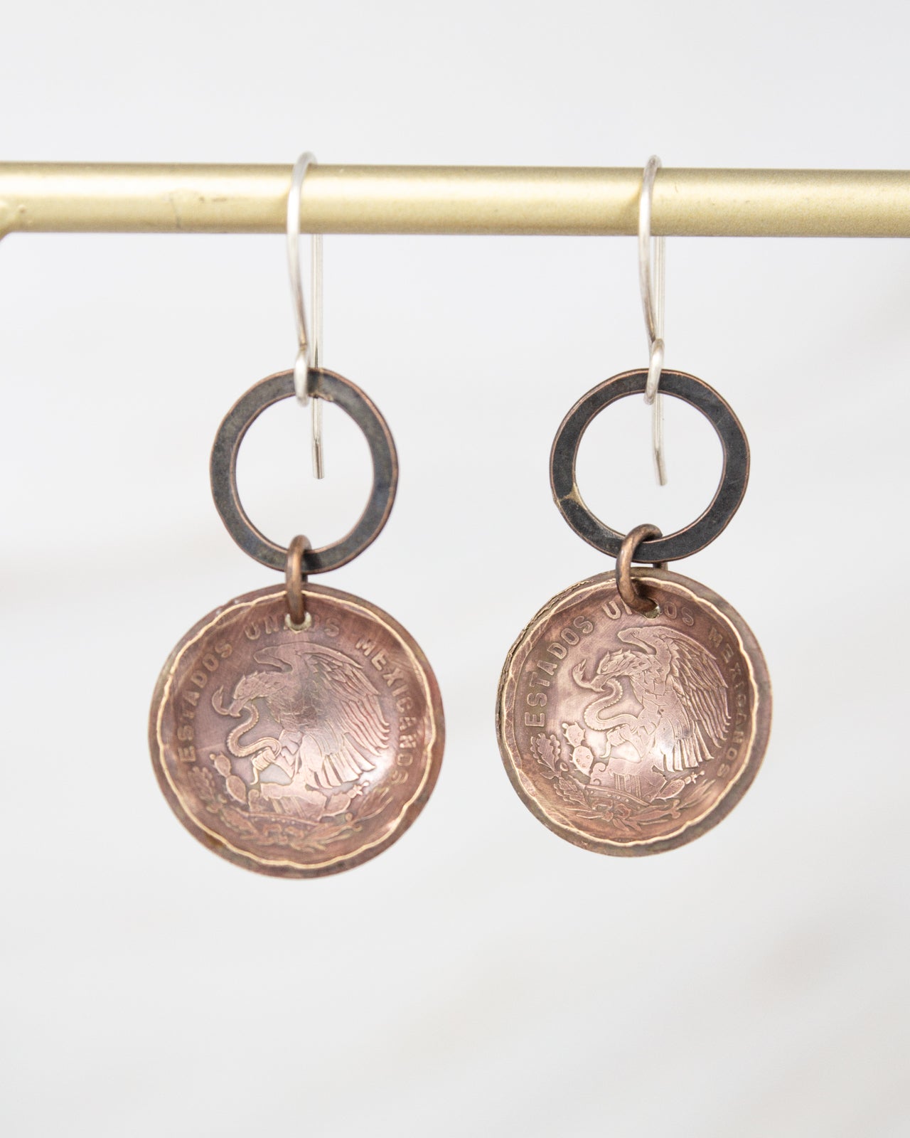 A pair of copper-colored earrings with a circular design featuring an image of a bird and the text "ESTADOS UNIDOS MEXICANOS" (Spanish for "United States of Mexico").