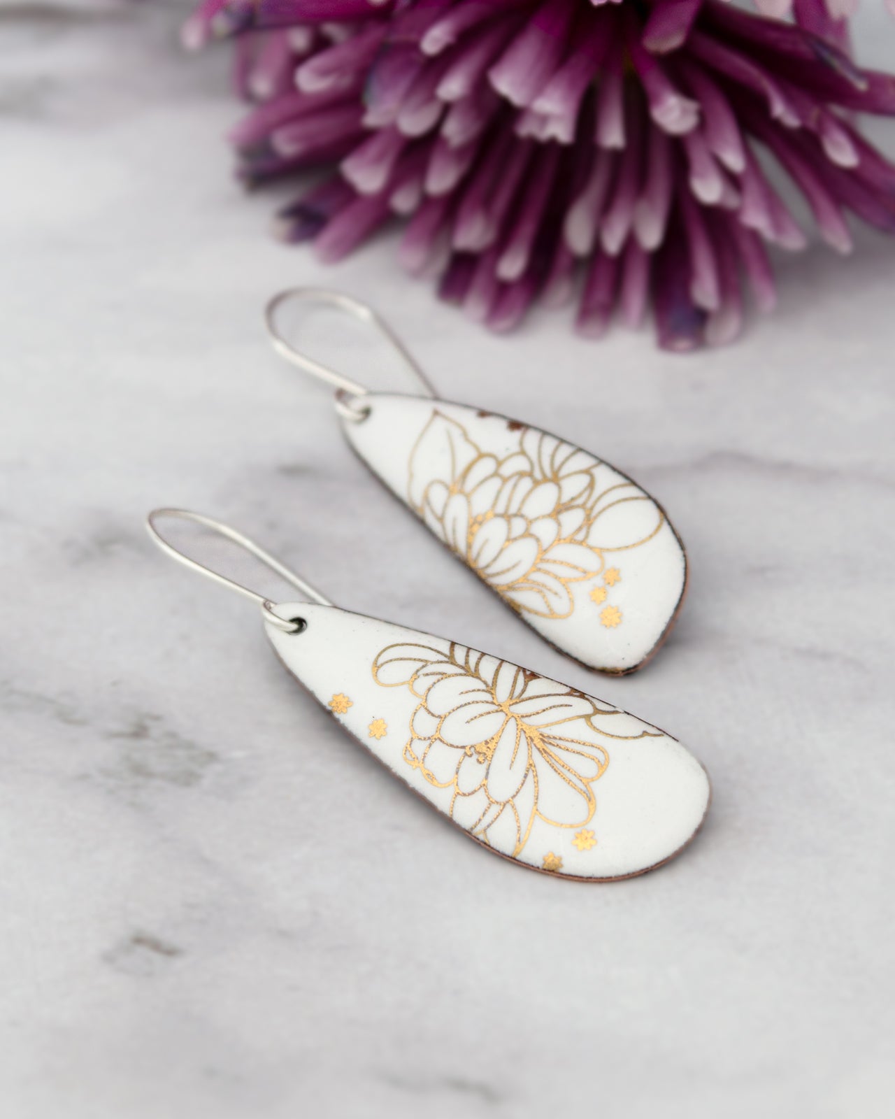 a pair of white earrings with gold floral designs, placed on a marble surface next to a purple flower.