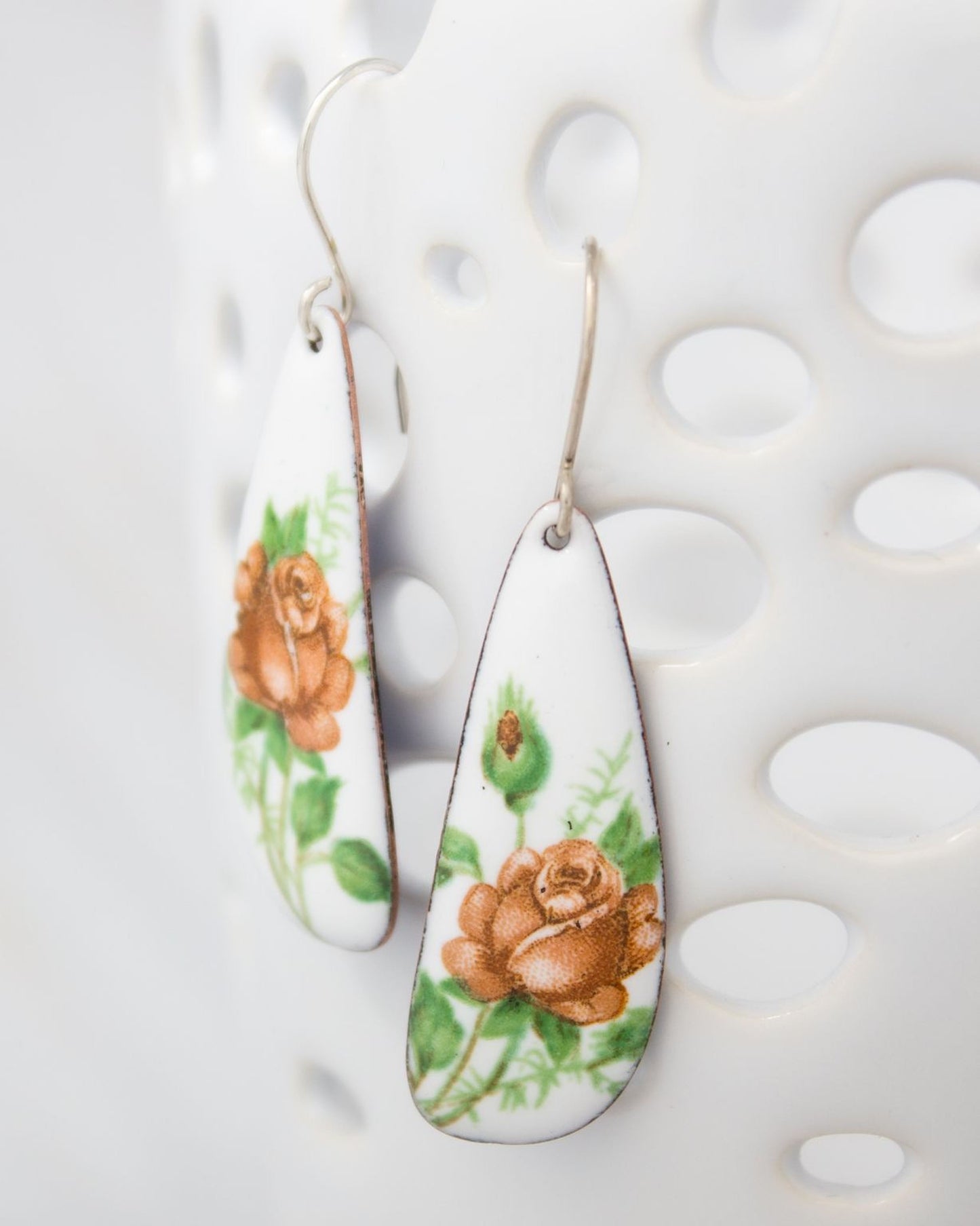A pair of white earrings with floral designs, featuring orange roses and green leaves.