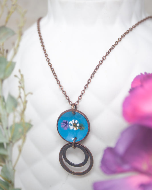 A necklace with a blue circular pendant featuring a floral design, hanging on a white mannequin.