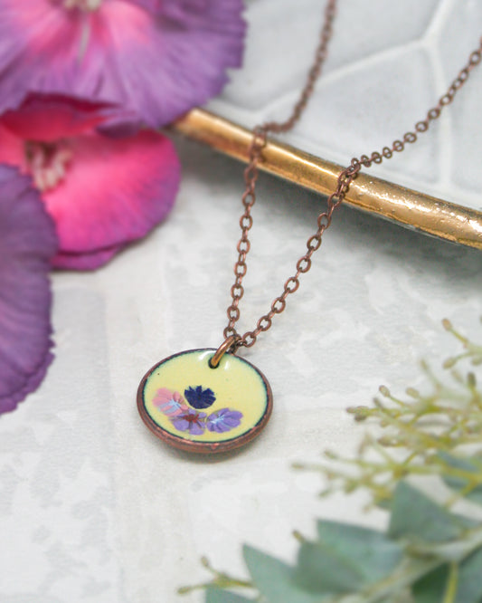 A necklace with a round pendant featuring a floral design rests on a white surface, surrounded by purple and green flowers.