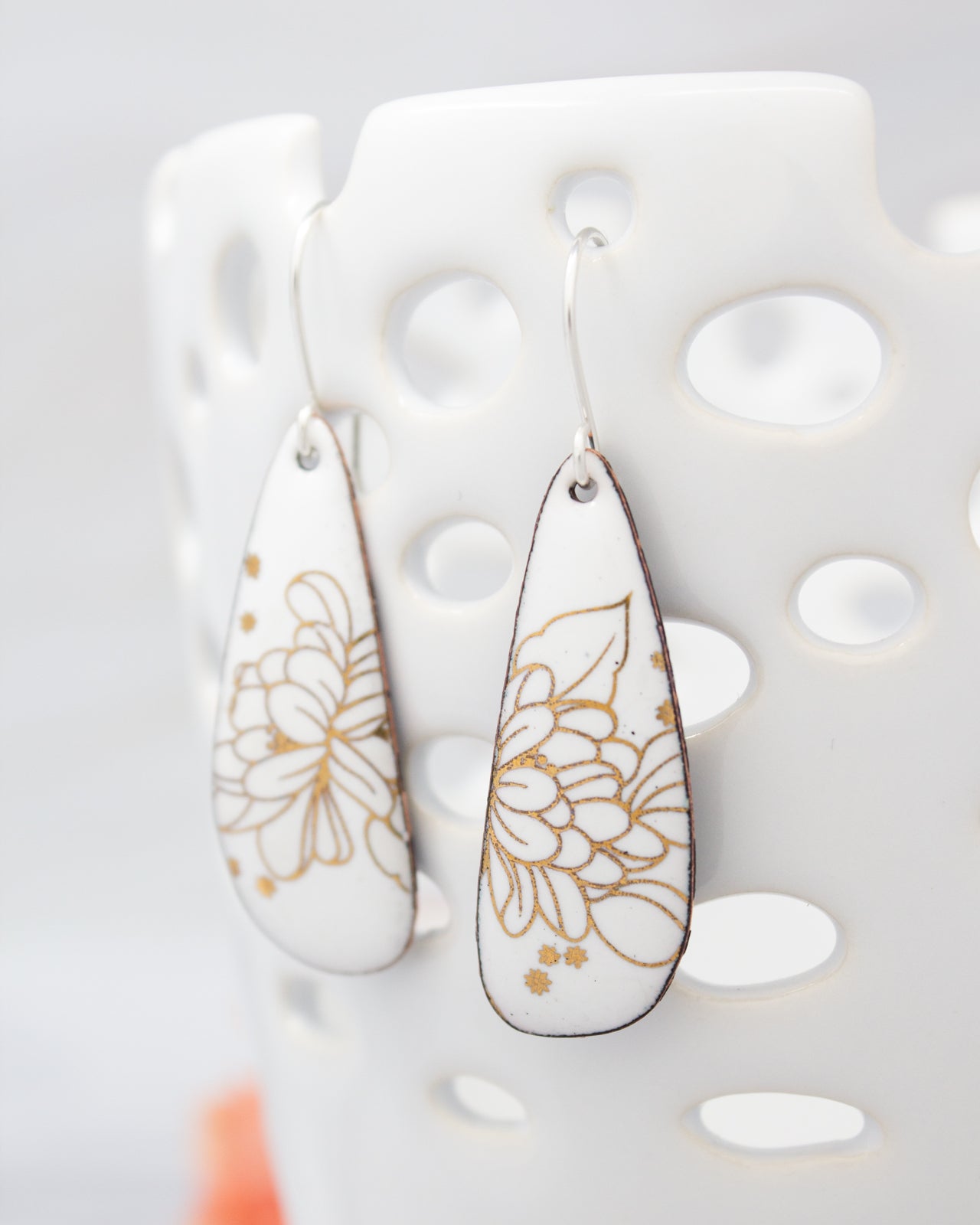 A pair of white earrings with gold floral designs on them, hanging from a white perforated holder.
