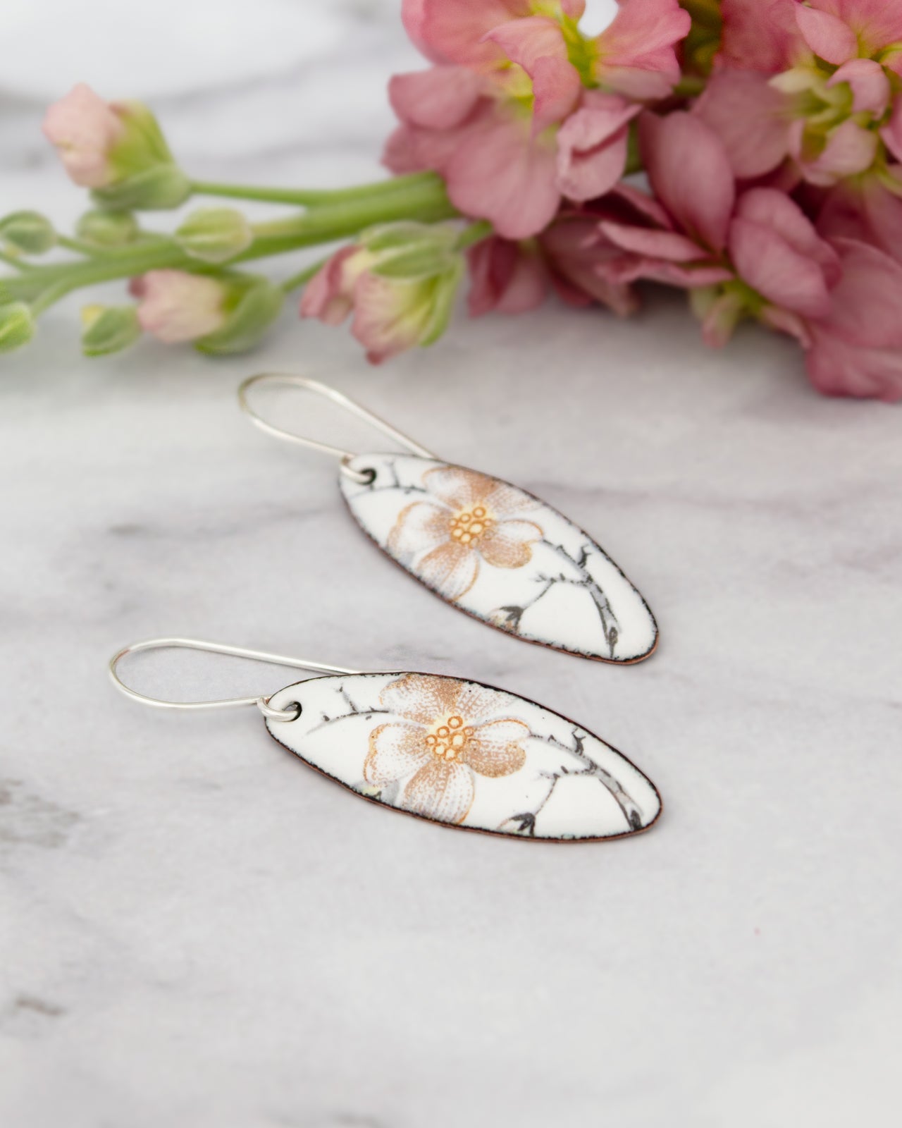 A pair of white earrings with a floral design rests on a marble surface, accompanied by a bouquet of pink flowers.