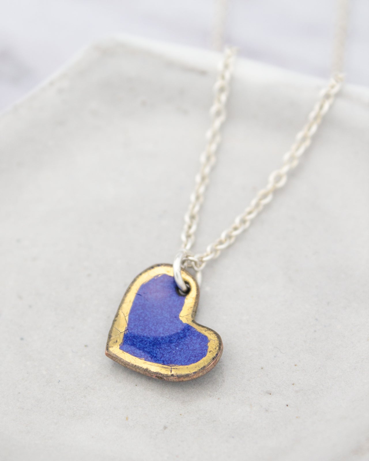 A delicate heart-shaped pendant with a blue background and a gold outline, hanging from a silver chain.