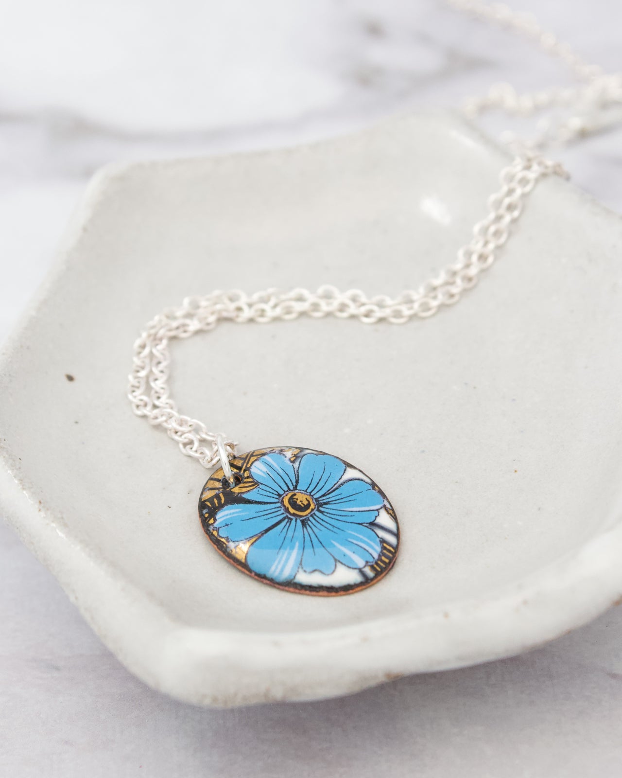 A necklace with a blue flower pendant is displayed on a white plate.