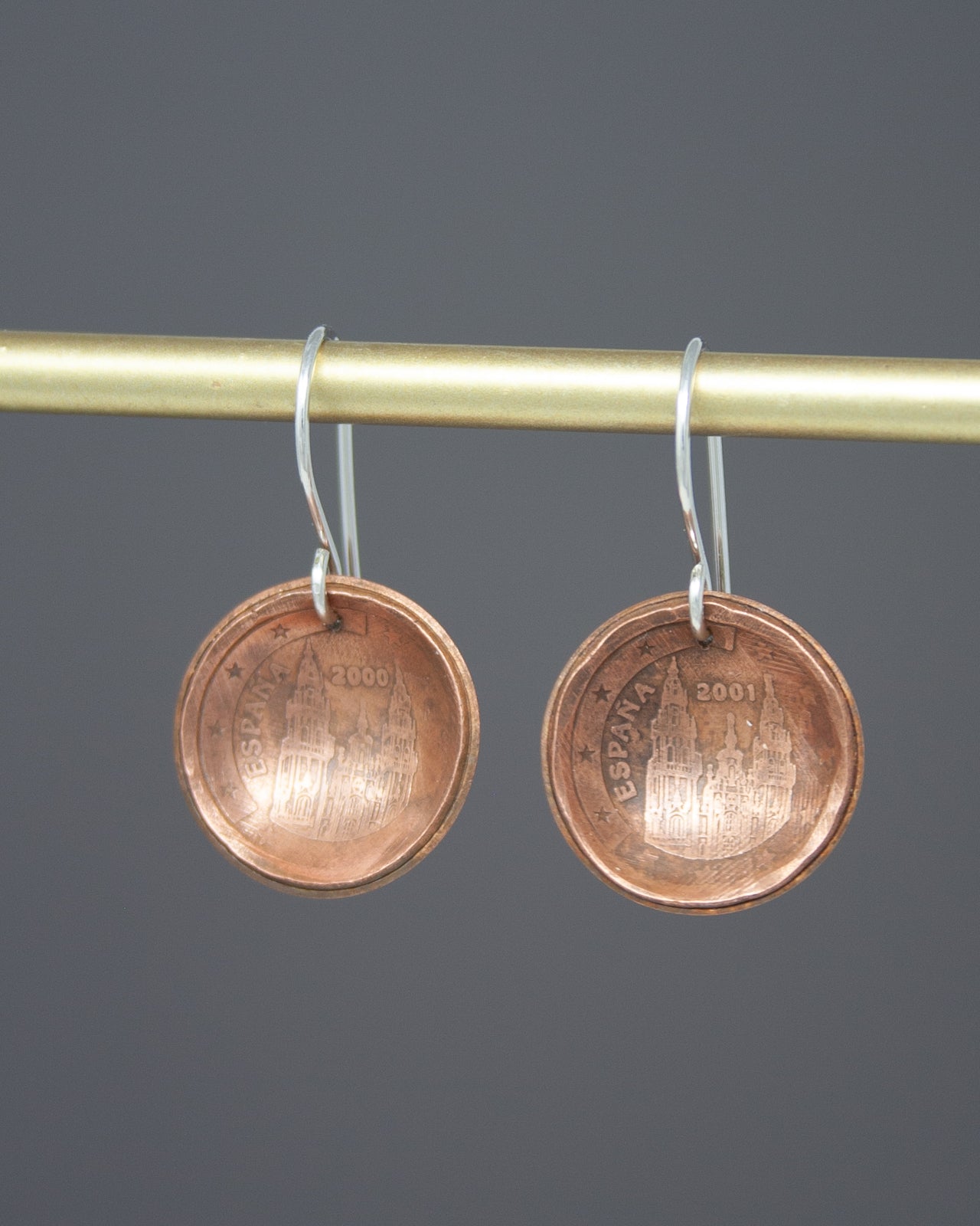 A pair of copper-colored earrings with a design of a building on them, hanging from a gold-colored metal bar.