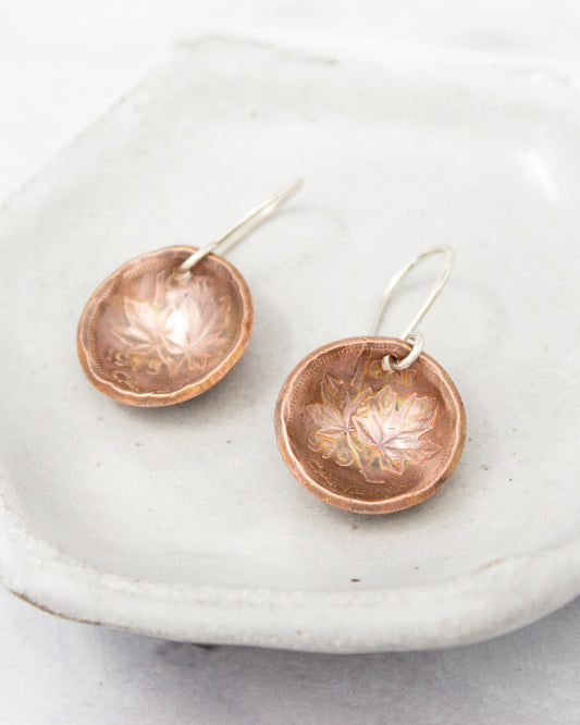 a pair of copper-colored earrings with leaf designs, displayed on a white plate.