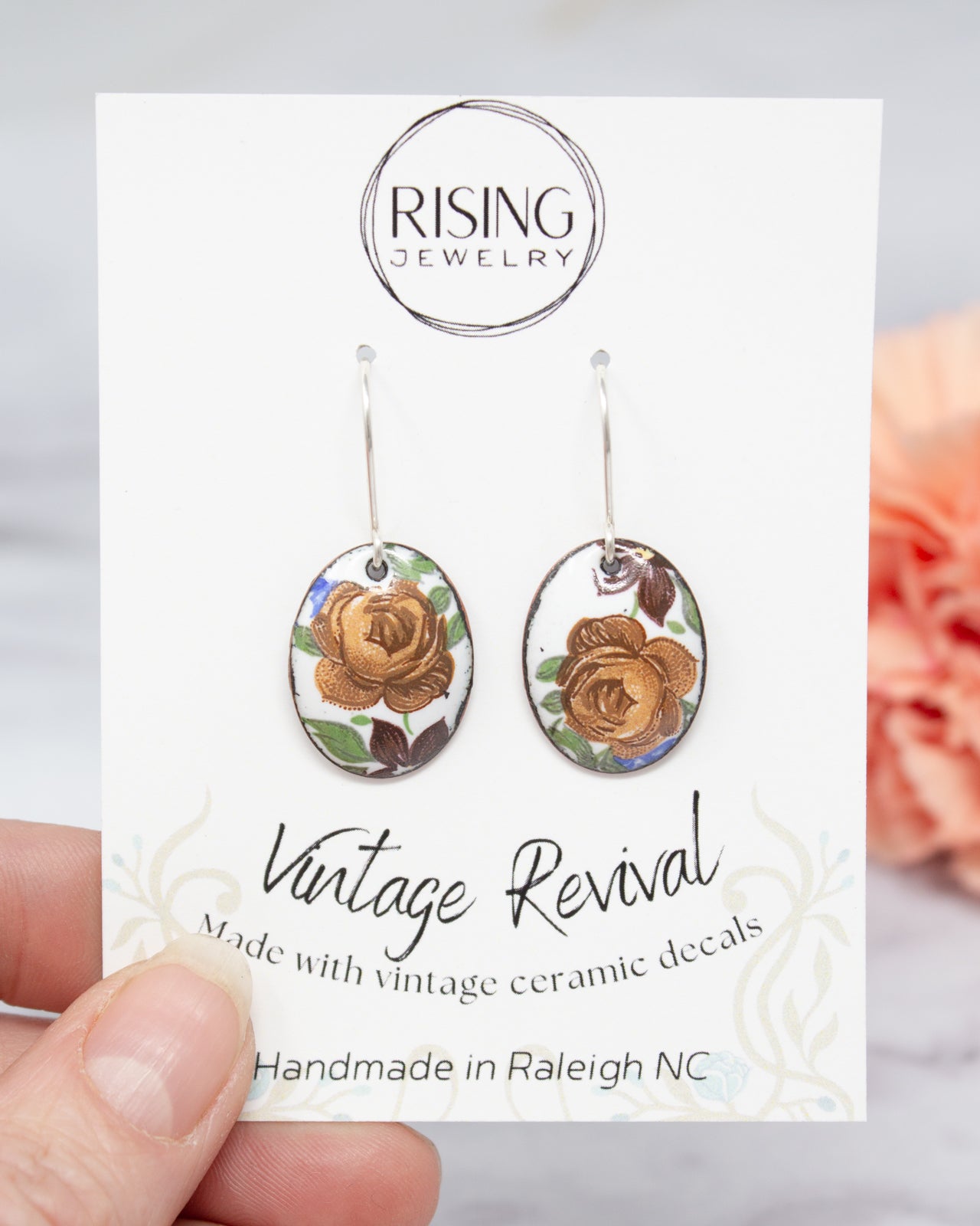 A hand holding a pair of earrings with a vintage-inspired design, featuring a ceramic rose and leaves. The earrings are described as "Vintage Revival" and "Handmade in Raleigh NC".