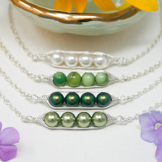 A collection of necklaces with various colored beads, including green, silver, and white pearls, arranged in a row. The necklaces are displayed on a white surface, with a bowl of yellow flowers and purple flowers in the background.