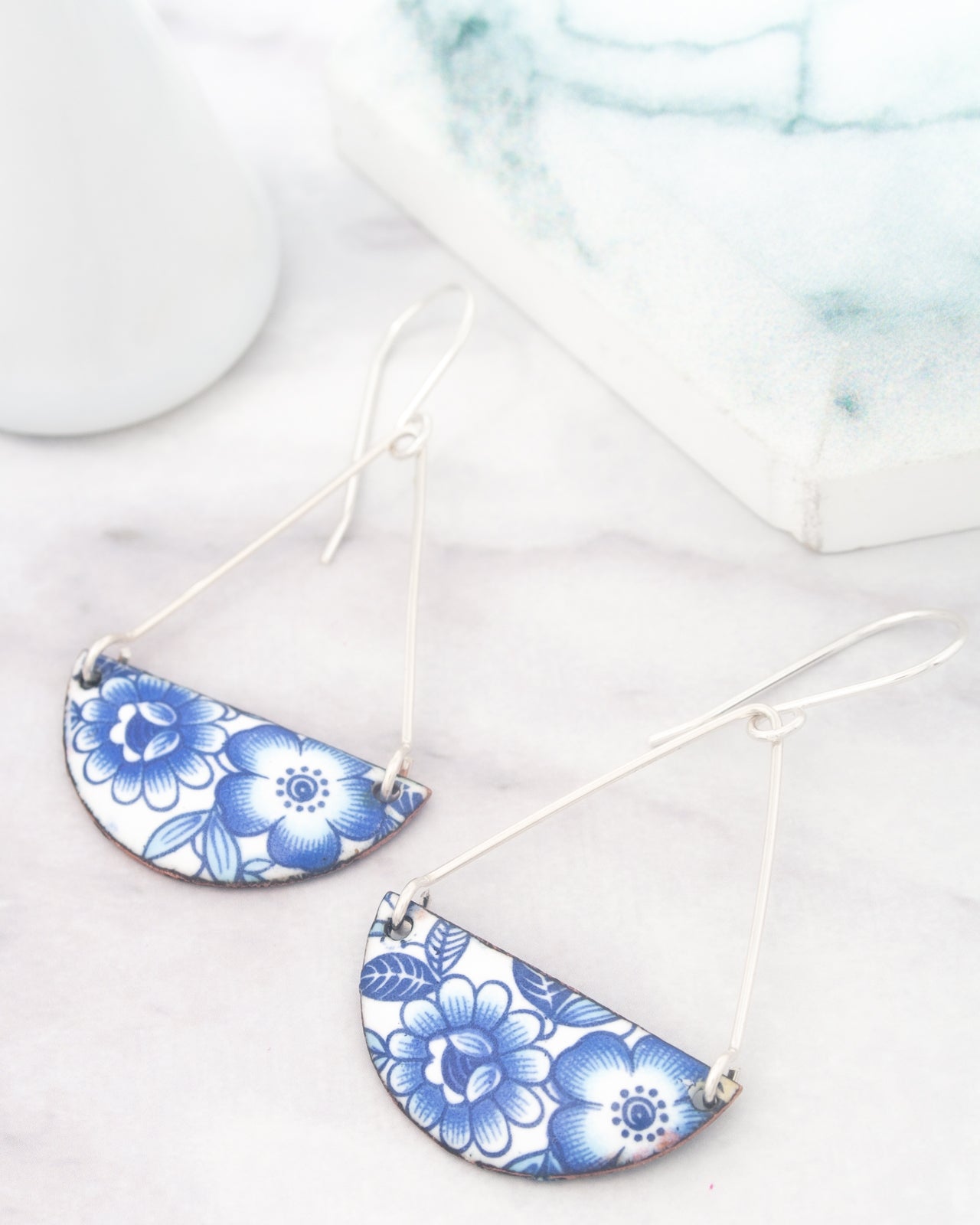 A pair of earrings with blue floral designs on a white marble surface, with a white vase and a white box in the background.