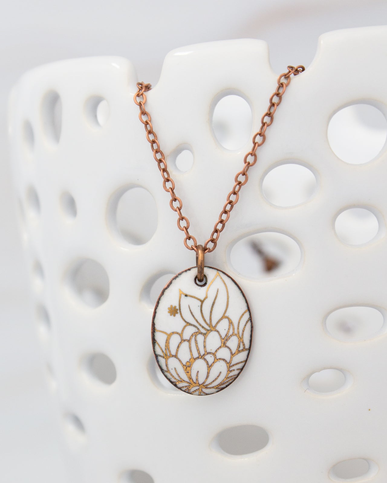 A white ceramic bowl with a copper-colored chain necklace featuring a white oval pendant with a gold floral design.