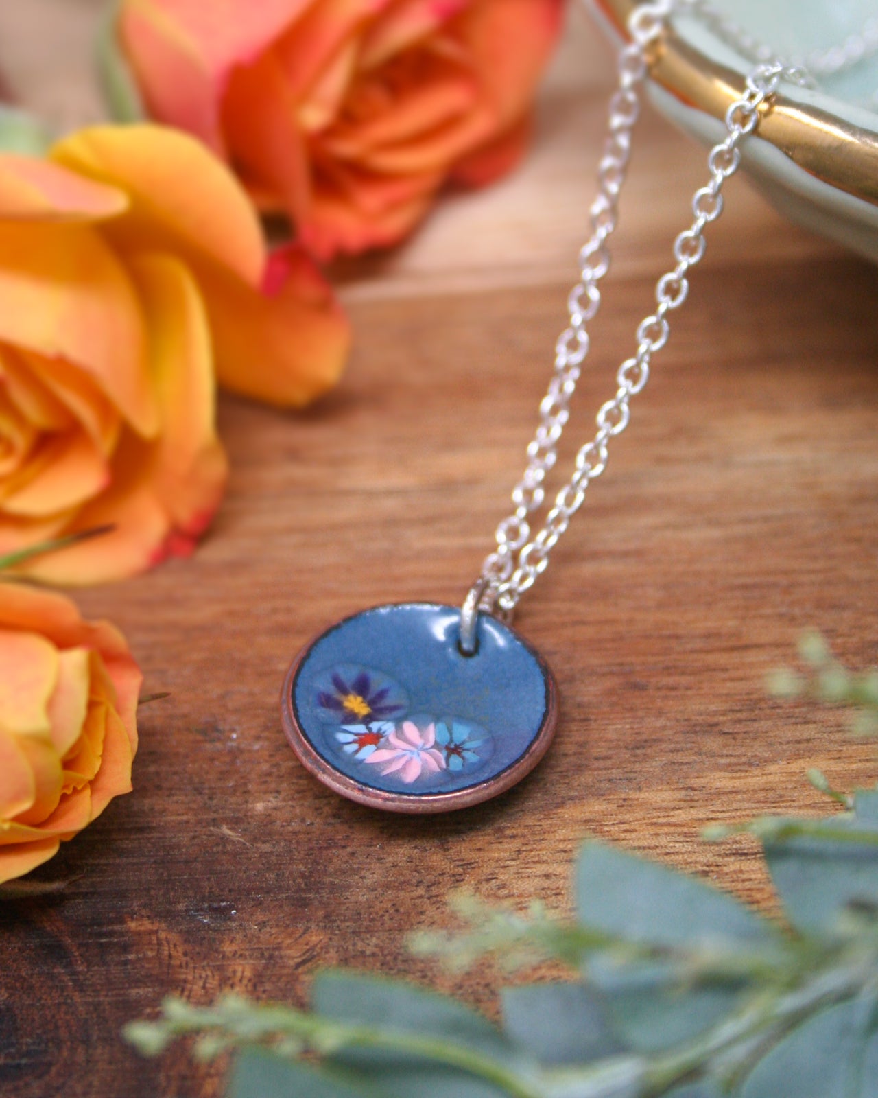 A blue pendant necklace with a floral design rests on a wooden surface, surrounded by vibrant orange and yellow roses.