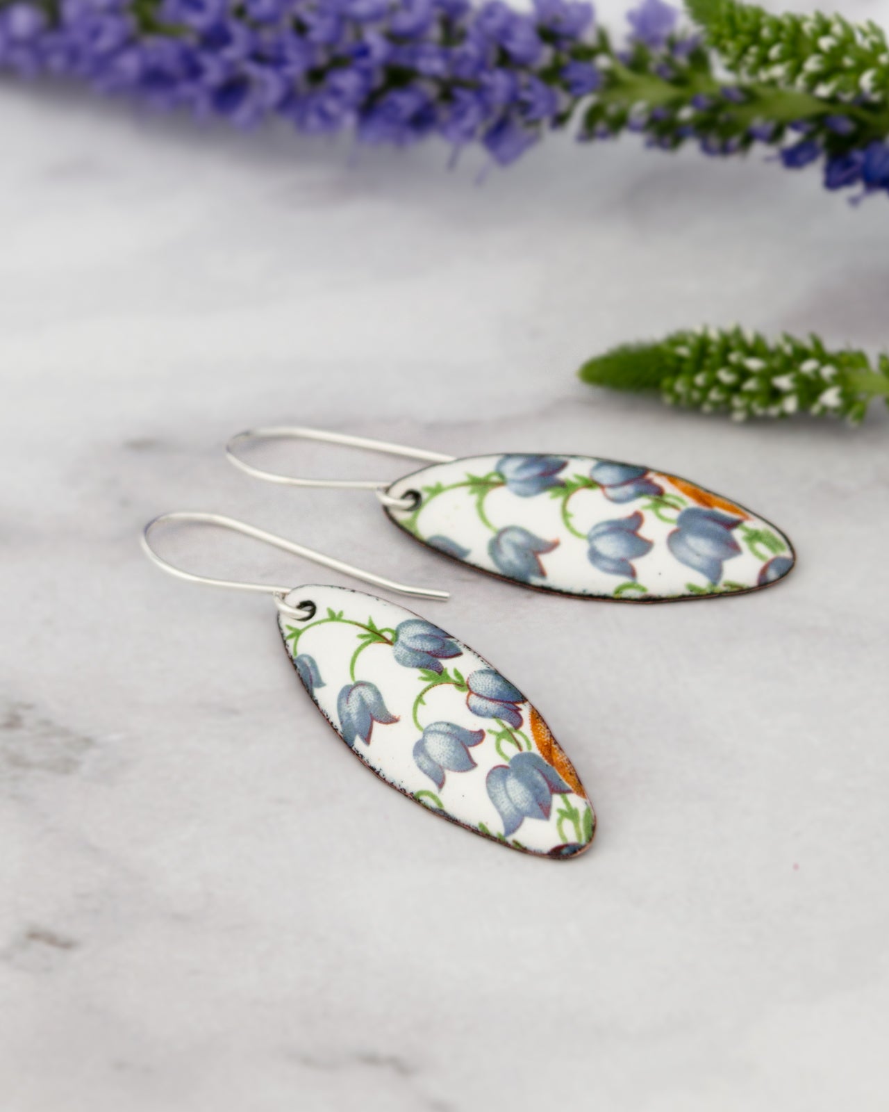 A pair of earrings with a floral pattern on a white background, with a sprig of lavender flowers and a green plant in the background.