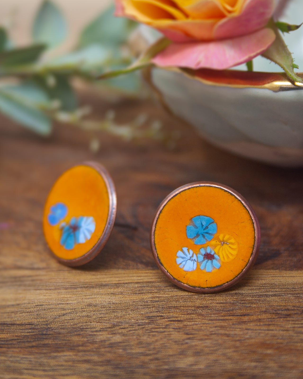 two round, orange-colored earrings with blue and yellow floral designs, placed on a wooden surface next to a white bowl with a pink rose inside.