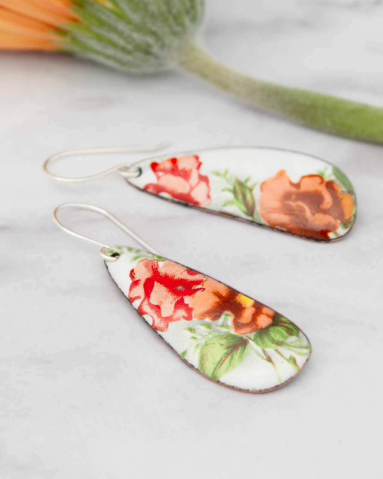 a pair of earrings with a floral design, including red and orange flowers, resting on a marble surface.