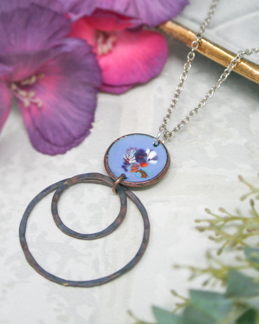 a necklace with a circular pendant, which appears to be a piece of jewelry. The pendant is surrounded by two large circular rings, creating a unique and eye-catching design. The necklace is placed on a white surface, with a few purple flowers visible in the background.
