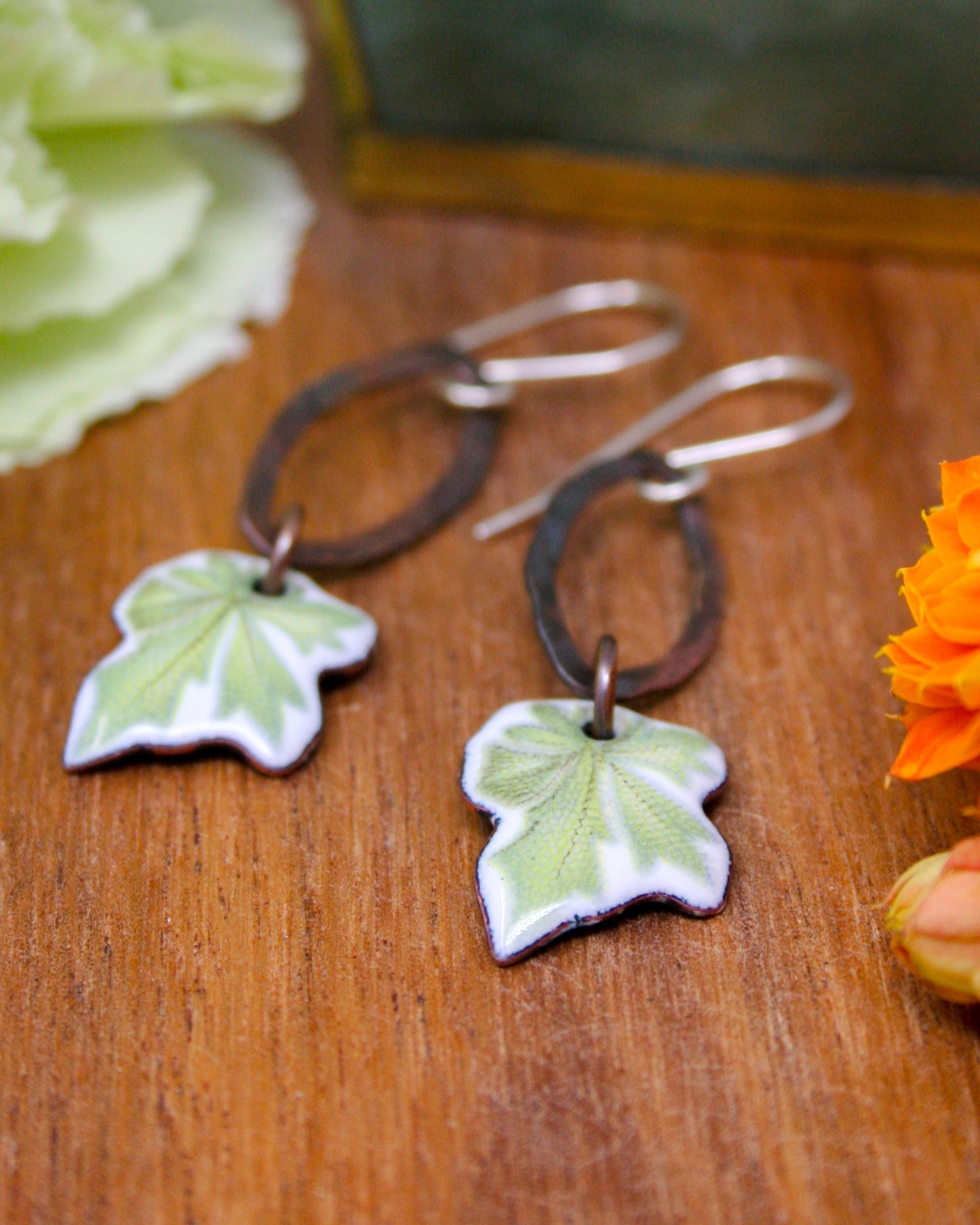 A pair of earrings with green and white leaf-shaped pendants, resting on a wooden surface alongside some flowers.