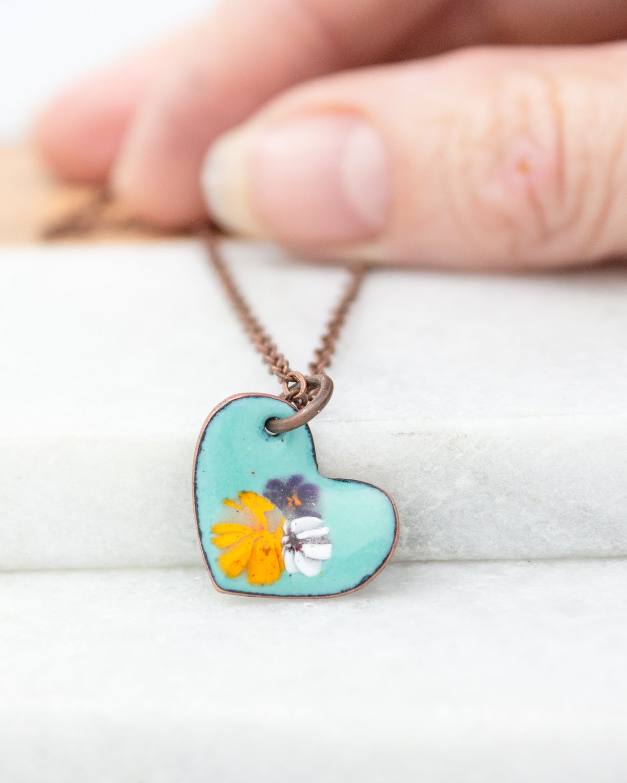 A hand is holding a small heart-shaped pendant necklace with a floral design.