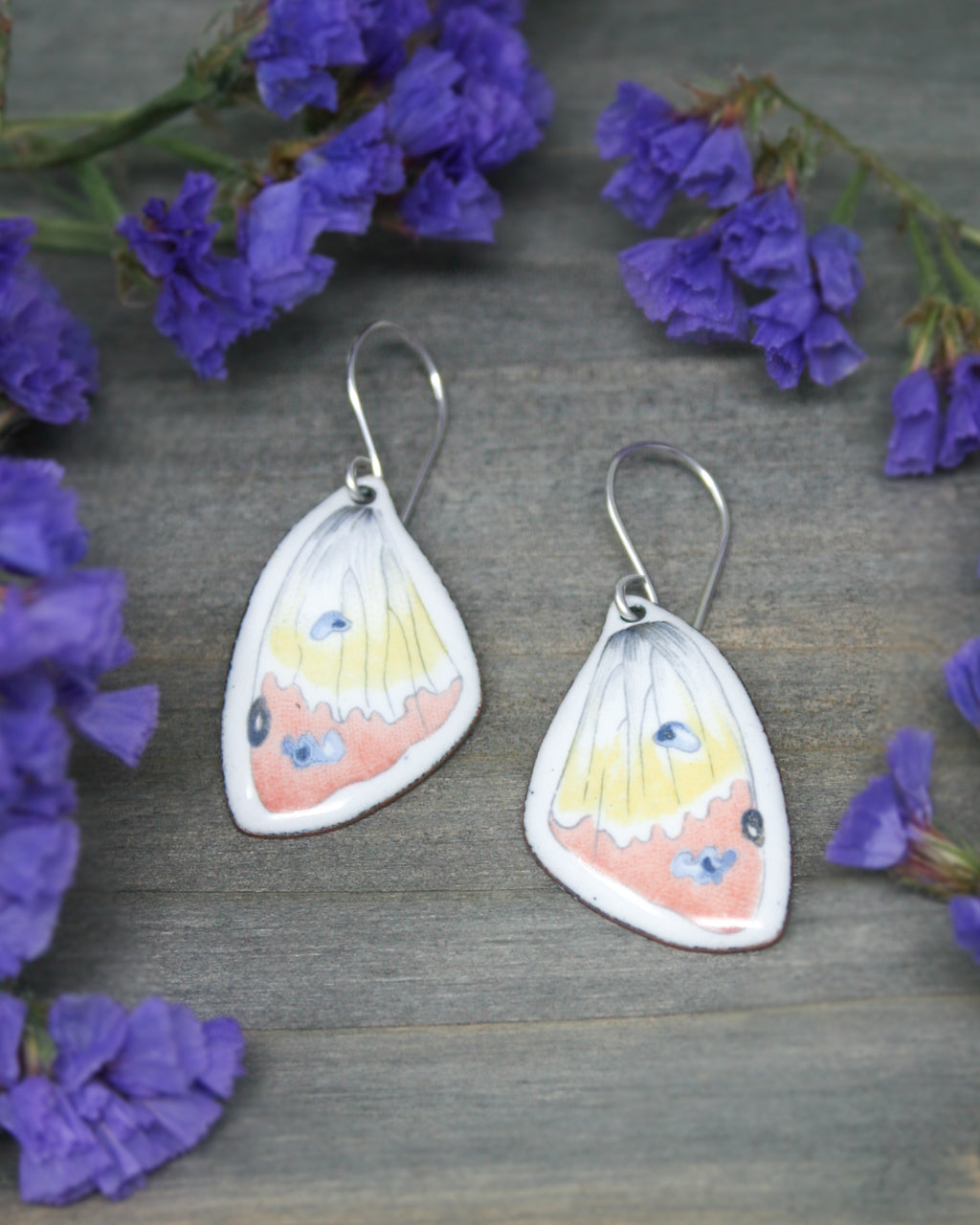 a pair of earrings with butterfly designs, placed on a wooden surface surrounded by purple flowers.
