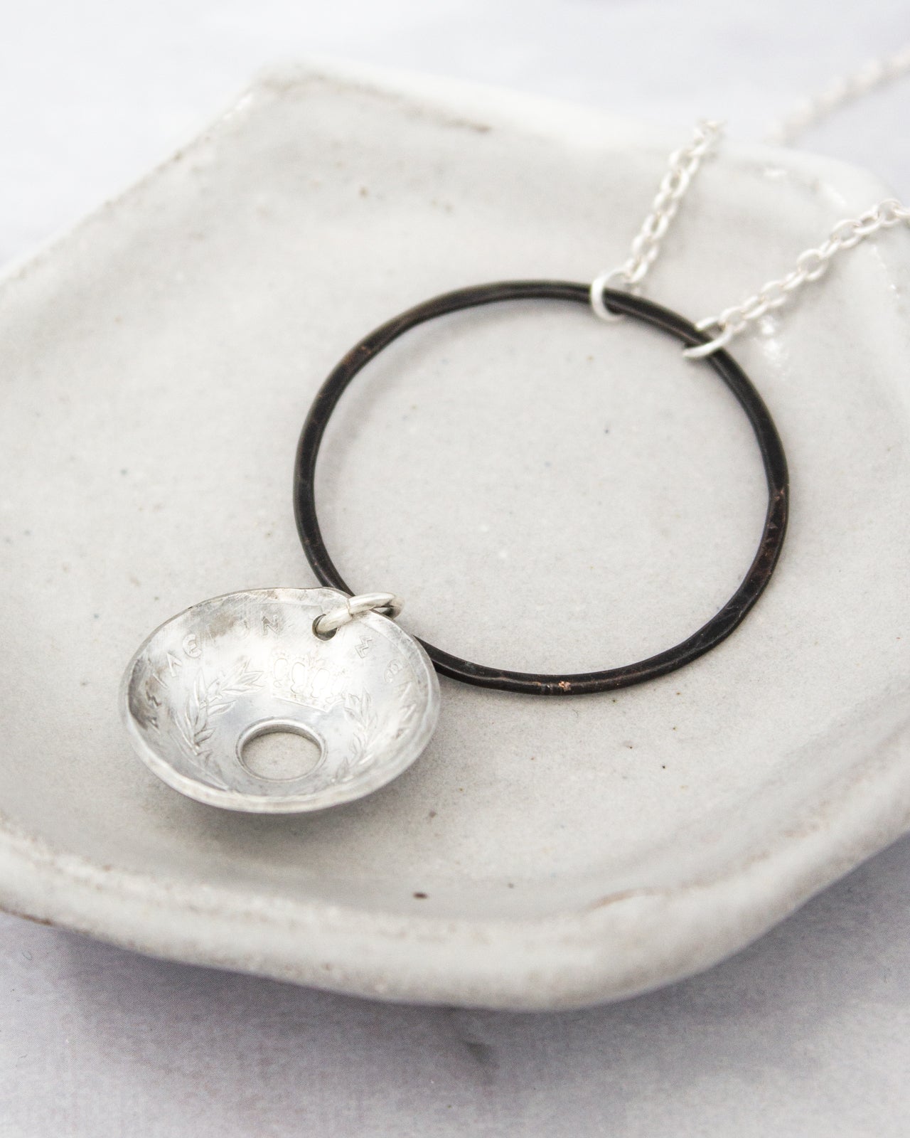 A silver necklace with a circular pendant and a small bowl-shaped charm hanging from it, resting on a white surface.